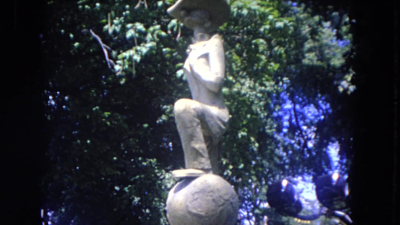 The 8mm camera lingers on the stone sculpture, with leafy trees and bright sky behind it.