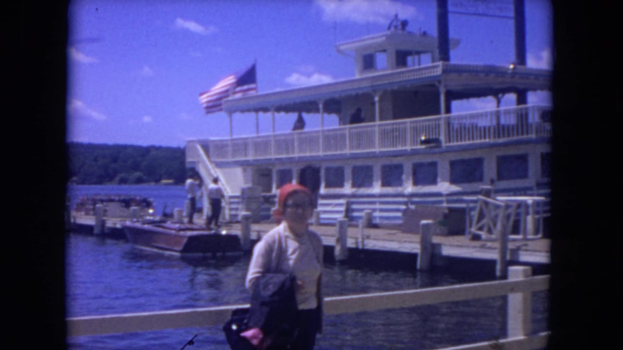 1972 Mississippi River frame showing a woman with a red headscarf near a white riverboat, dock railings, water, trees, and an American flag.