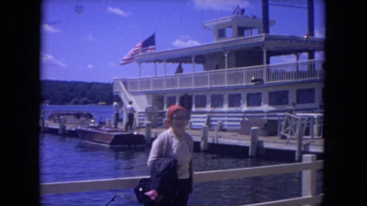 Woman in a red headscarf stands on a dock in front of a white riverboat and American flag.
