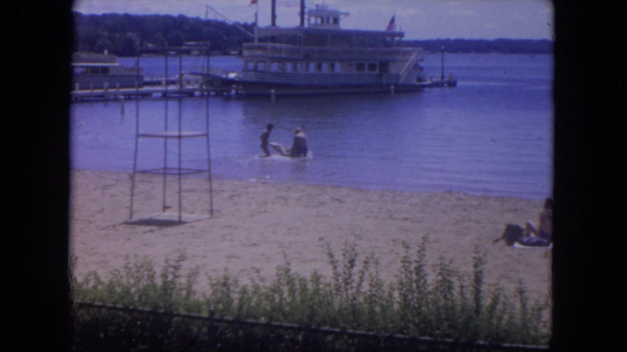 The clip cuts to a Mississippi River beach scene with people in shallow water and a white passenger boat docked across the riverfront.