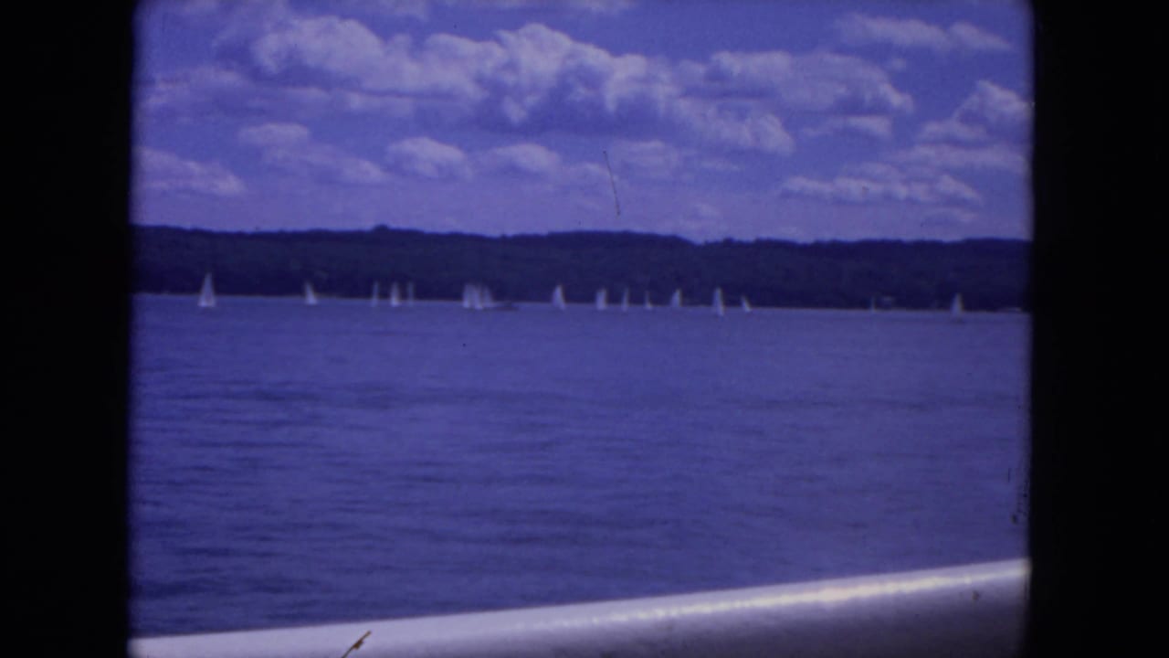 Wide water view with small sailboats near a wooded shoreline under scattered clouds.