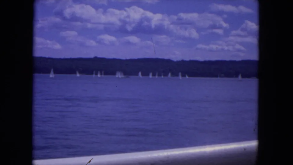 1972 8mm view of the Mississippi River with distant sailboats, blue water, wooded shore, and a cloud-filled sky.