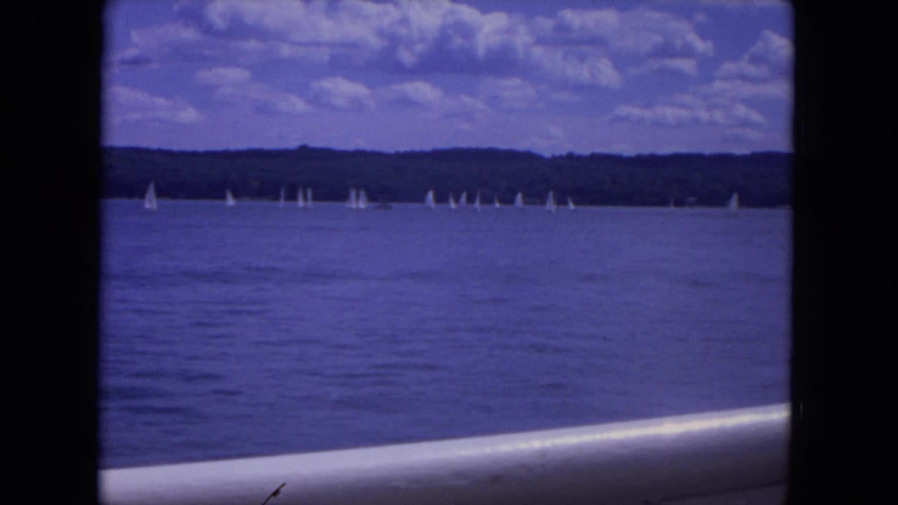 Wide 1972 Mississippi River shot with scattered white sailboats, a dark wooded shoreline, and blue-purple 8mm color tone.