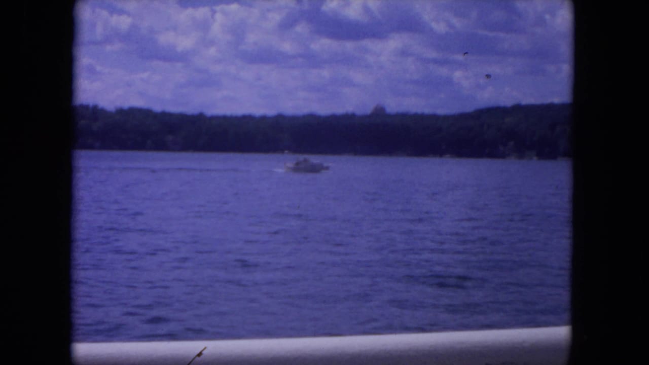 1972 8mm river scene showing a small speedboat on blue water, distant trees, and a cloudy summer sky.