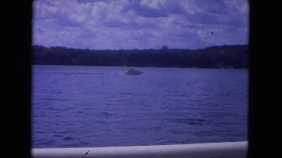 1972 8mm river scene showing a small speedboat on blue water, distant trees, and a cloudy summer sky.
