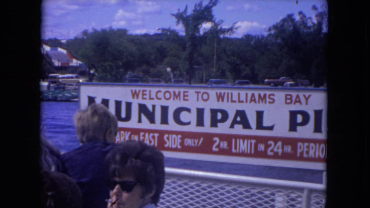 1972 8mm deck view approaching a Williams Bay Municipal Pier sign, with passengers and parked cars visible beyond.