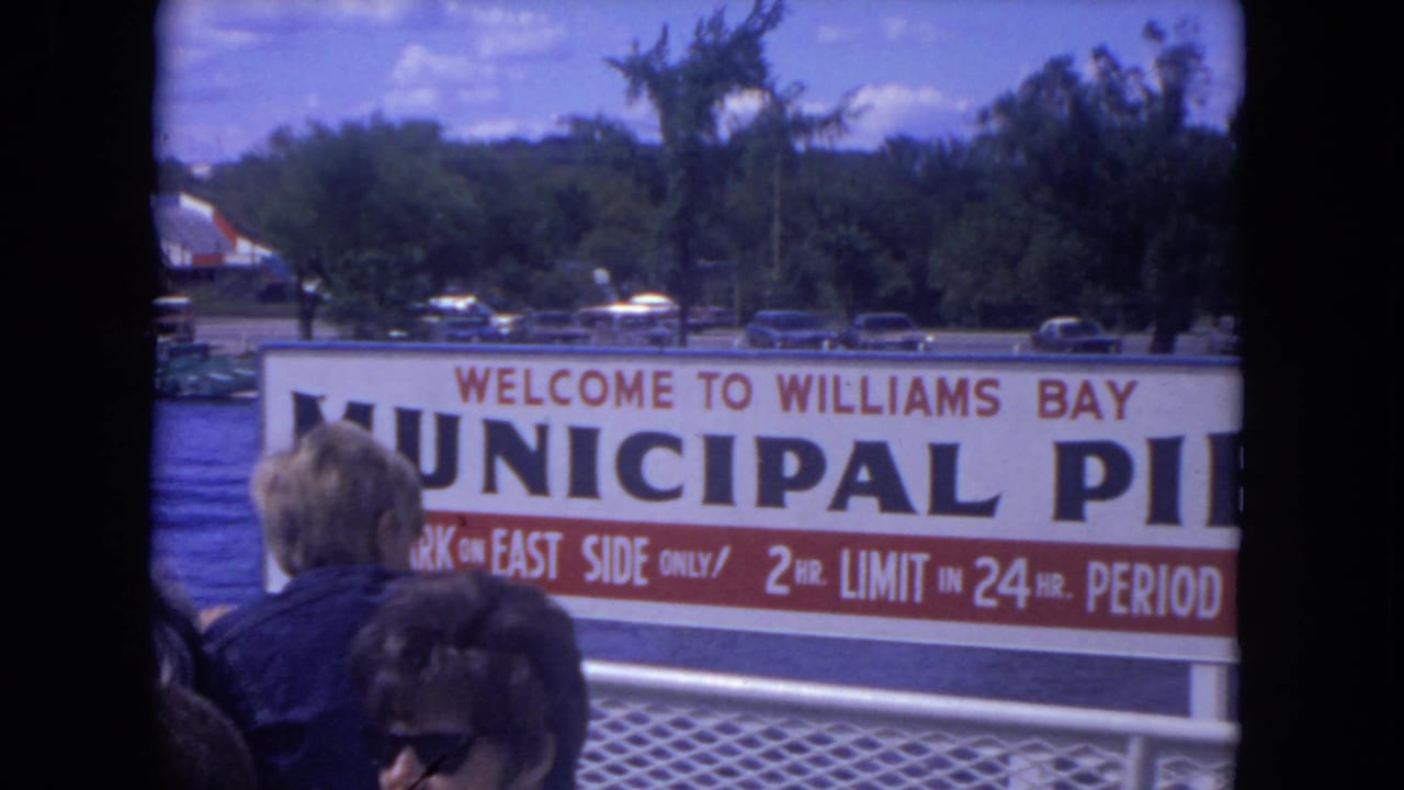 1972 8mm view of the Williams Bay Municipal Pier sign, deck passengers, trees, and cars near the waterfront.