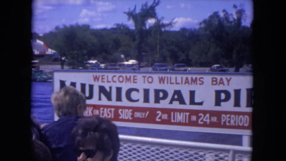 1972 8mm view of the Williams Bay Municipal Pier sign, deck passengers, trees, and cars near the waterfront.