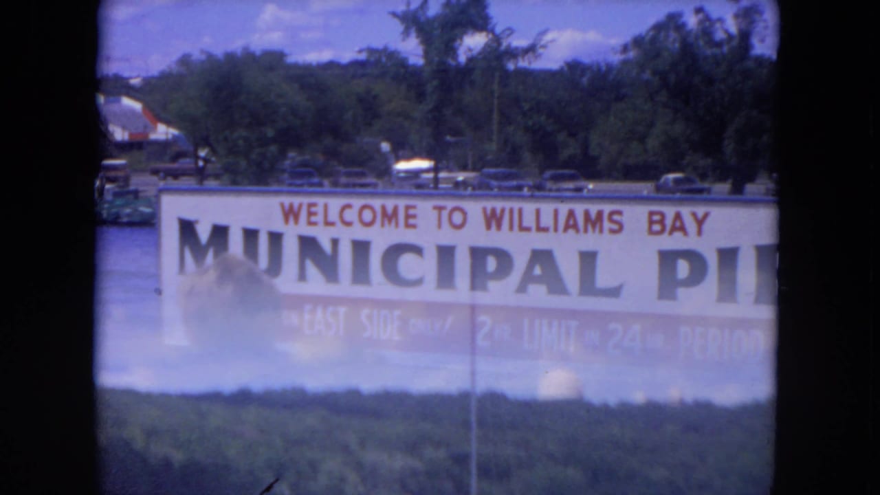 1972 8mm frame with the Williams Bay Municipal Pier sign partly obscured by reflection or exposure, with shoreline and trees beyond.