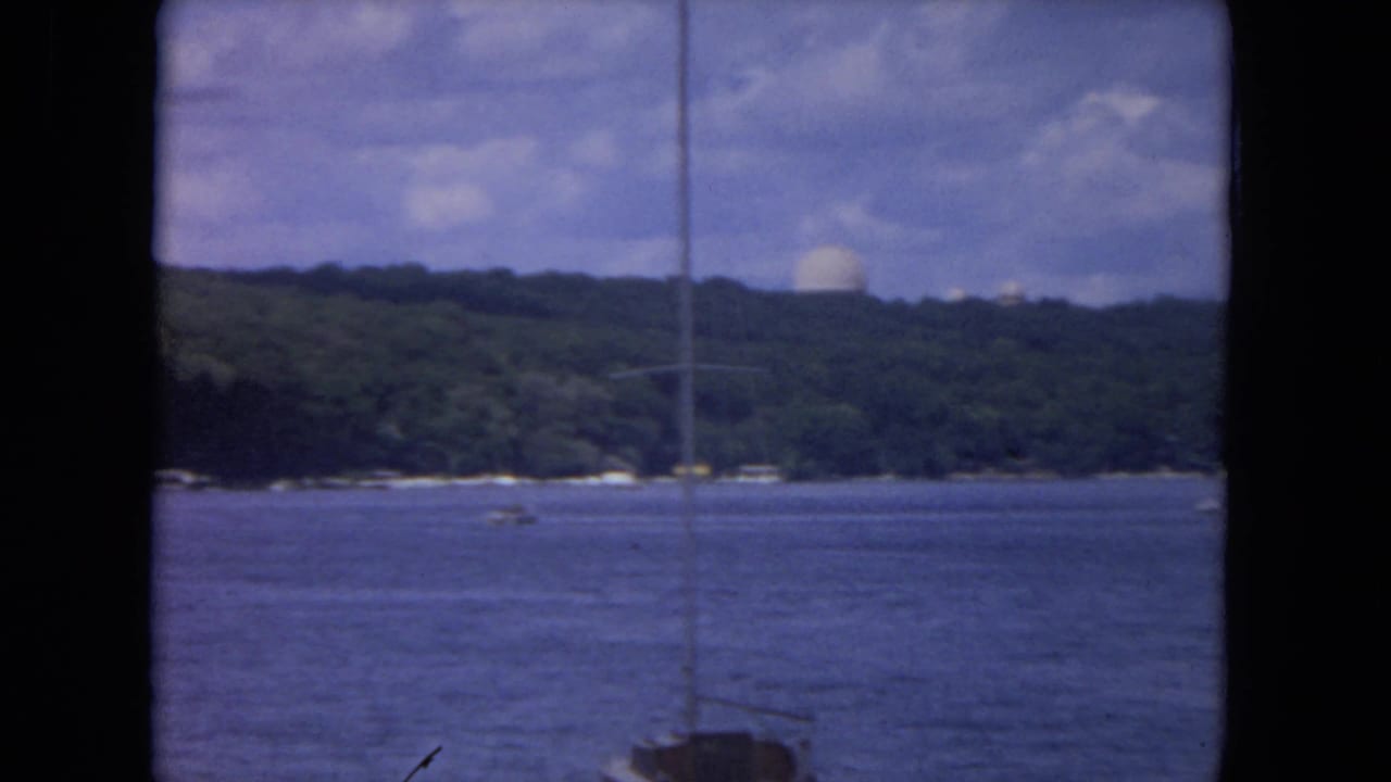 Sailboat mast in front of calm river water and wooded shoreline under a cloudy sky.