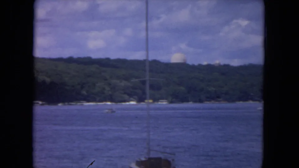 1972 8mm view of the Mississippi River with a sailboat mast crossing calm water, wooded hills, and a distant white dome.