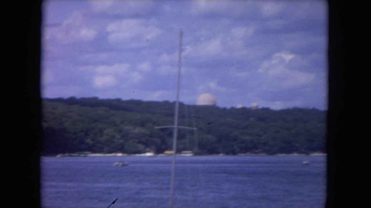 River view with a tall sailboat mast, wooded shore, small boats, and a distant white dome.