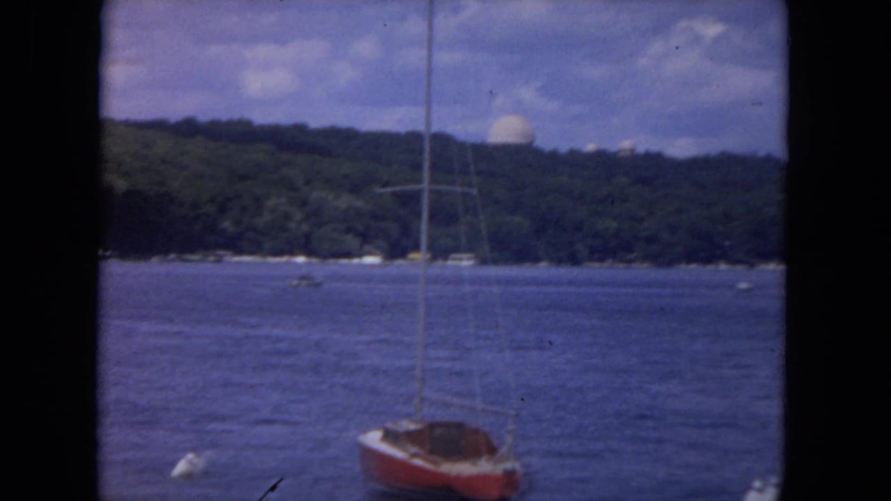 1972 river boating frame with a red-hulled sailboat in the foreground and the wooded Mississippi River shoreline beyond.