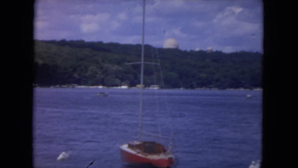1972 river boating frame with a red-hulled sailboat in the foreground and the wooded Mississippi River shoreline beyond.