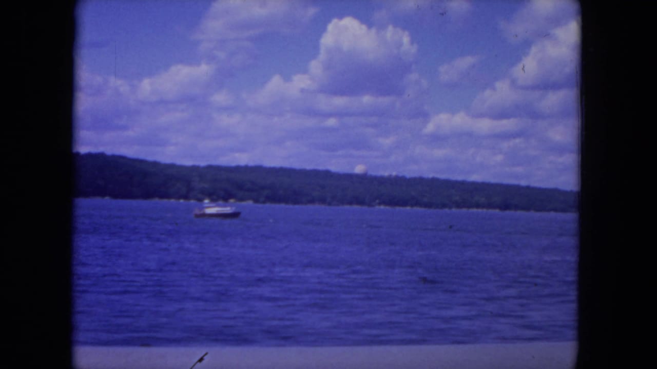 1972 8mm view of a small motorboat crossing blue water on the Mississippi River with a distant tree-lined shore under clouds.