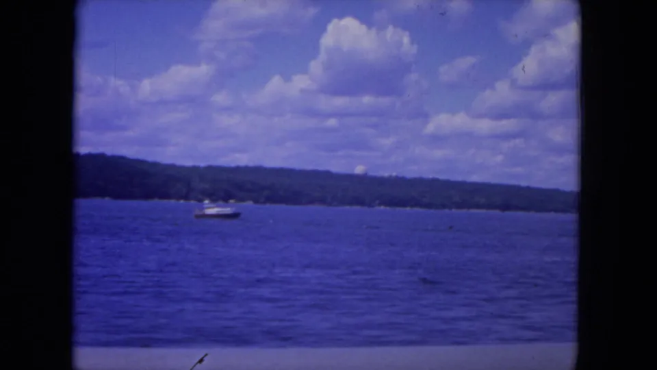 1972 8mm view of a small motorboat crossing blue water on the Mississippi River with a distant tree-lined shore under clouds.