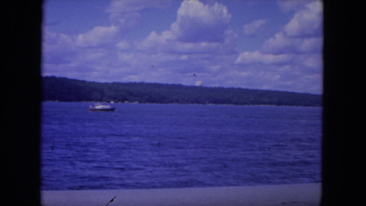 Small motorboat on the Mississippi River, framed by blue water, a wooded horizon, and a partly cloudy summer sky in 1972 home-movie footage.
