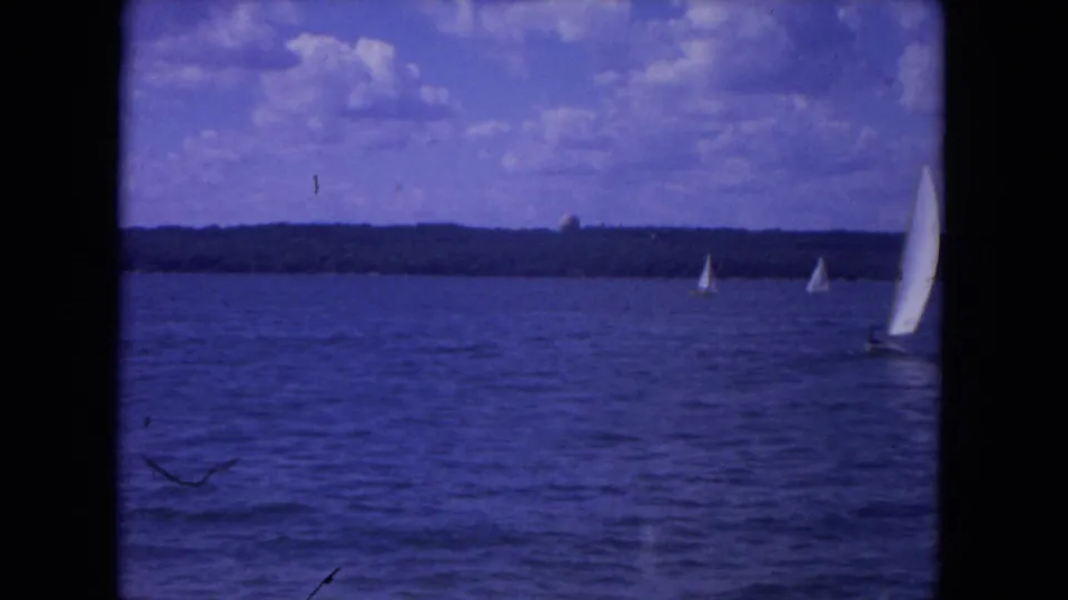 1972 8mm Mississippi River scene with white sailboats spread across blue water against a distant tree-lined shore and cloudy sky.
