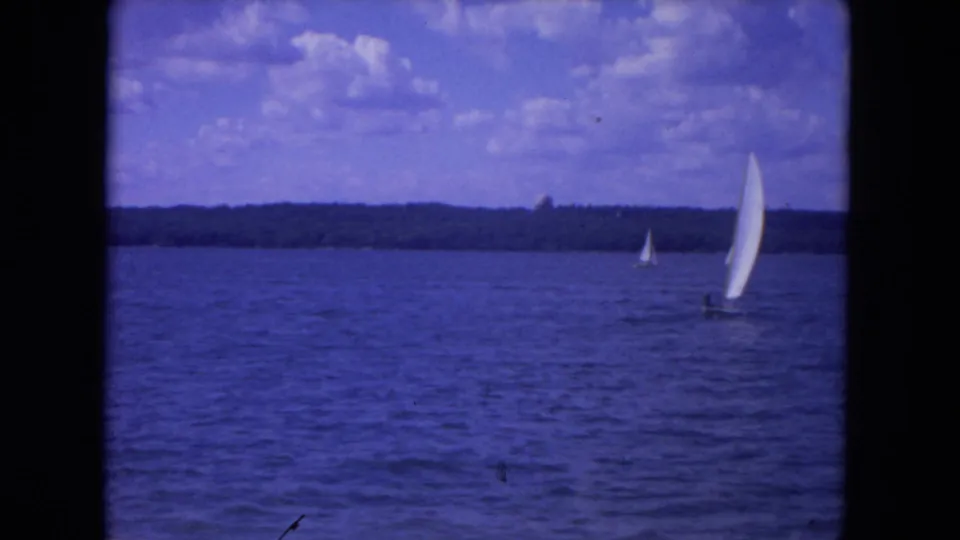 A white sailboat crosses the Mississippi River in 1972 home-movie footage, with open water, clouds, and a wooded horizon behind it.