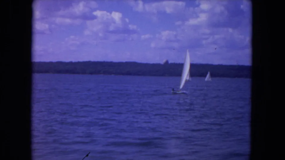 Sailboats cross the Mississippi River in a blue-toned 8mm frame with trees and clouds beyond the water.