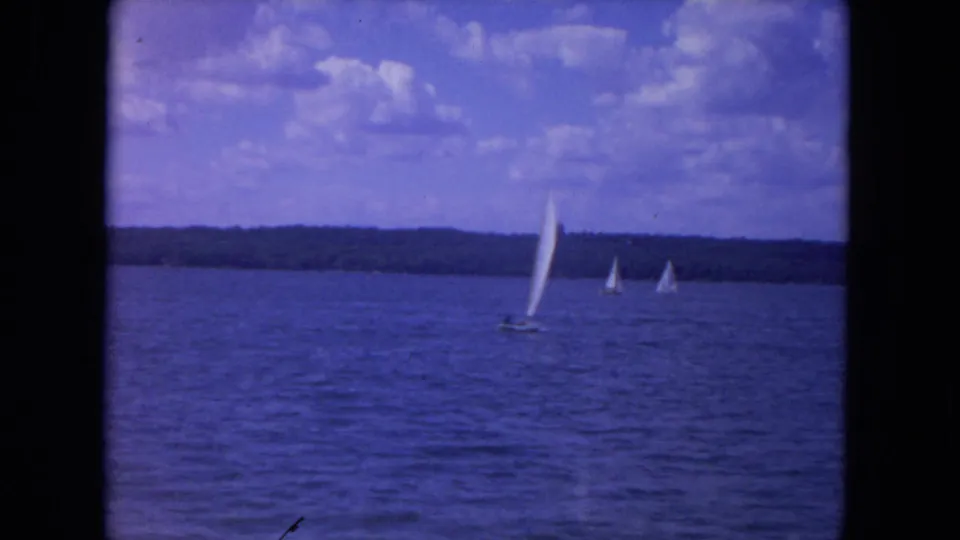 Home-movie footage shows a white sailboat on the Mississippi River, with distant boats and a wooded horizon.