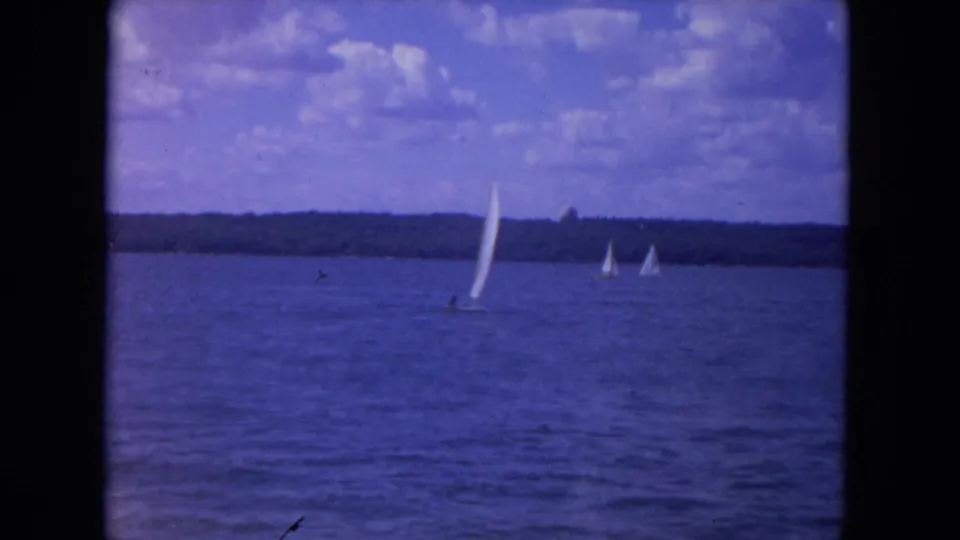 Blue-cast 8mm frame of sailboats on the Mississippi River, with open water, clouds, and a far tree line.