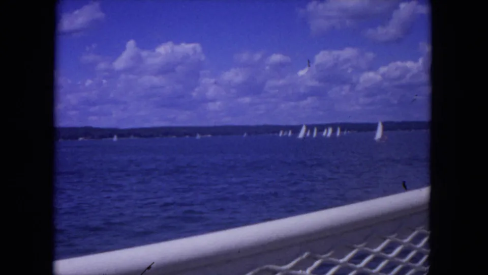 Wide 1972 home-movie frame of Mississippi River sailboats, blue water, cloudy sky, and foreground railing.