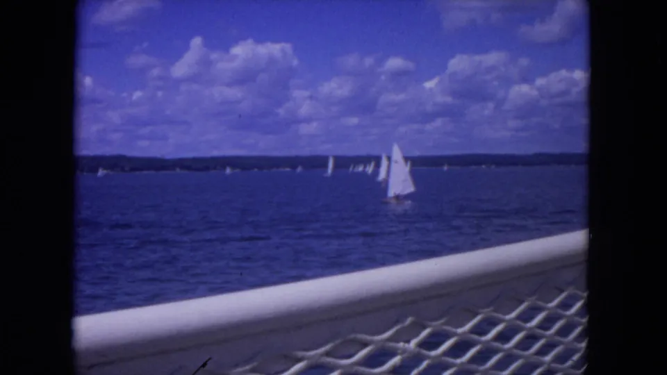 1972 Mississippi River home-movie frame showing a closer white sailboat and distant regatta sails beyond the boat railing.
