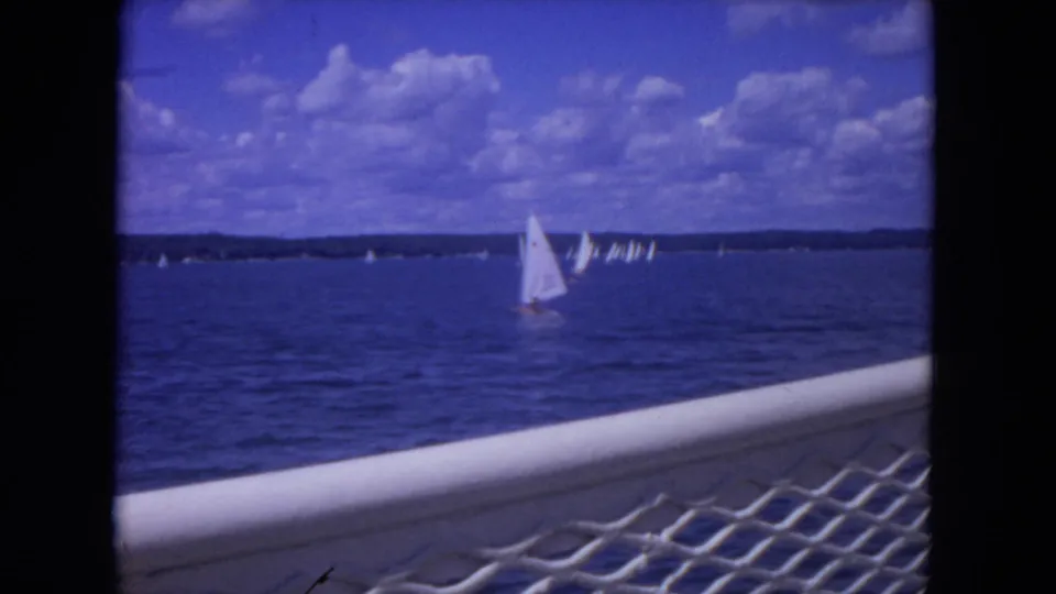 View from a moving vessel on the Mississippi River, with white sailboats under scattered clouds and a railing in the foreground.