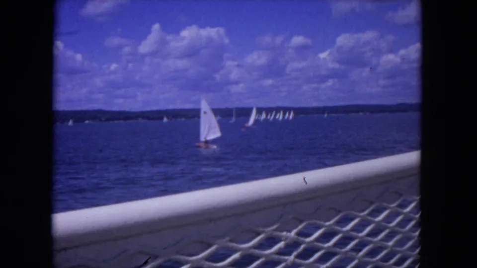 Home-movie frame showing a small sailboat on river water, distant clustered sails, shoreline, and foreground boat railing.