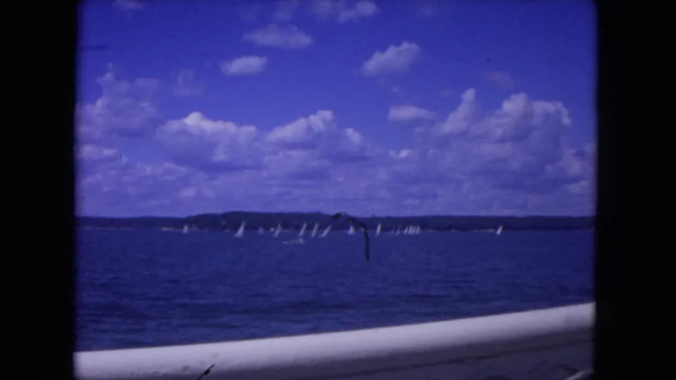 Wide 1972 Mississippi River view with multiple distant sailboats, open water, low shoreline, clouds, and a vessel rail at the bottom.