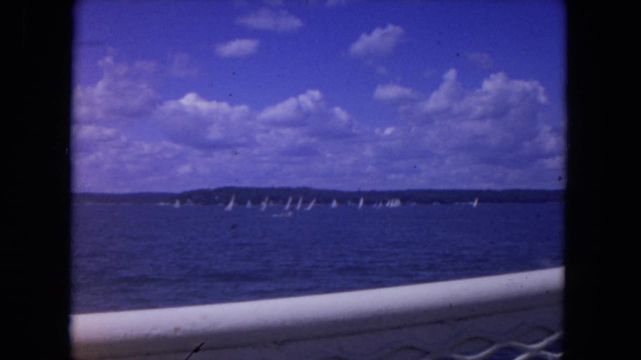 Distant sailboats on open blue water beneath scattered clouds, seen past a boat railing.