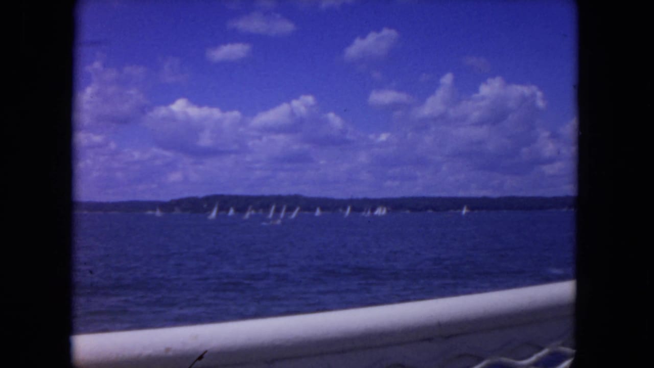 Sailboats spread across the horizon on blue water, with a boat railing in the foreground.
