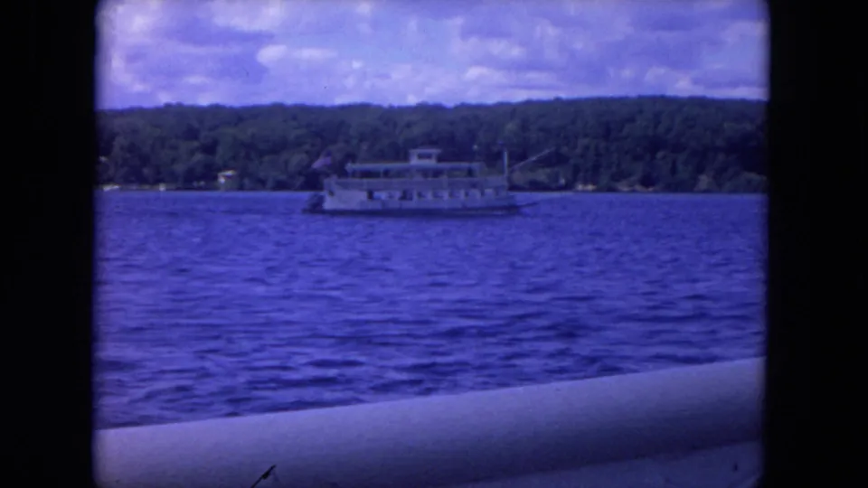 1972 8mm footage shows a paddlewheel-style riverboat moving across blue water with a tree-lined shore behind it.