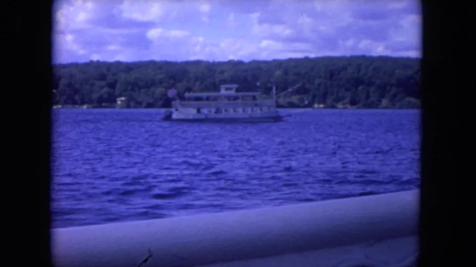 A paddlewheel-style riverboat continues past a tree-lined Mississippi River shore in blue-shifted 1972 8mm home-movie footage.