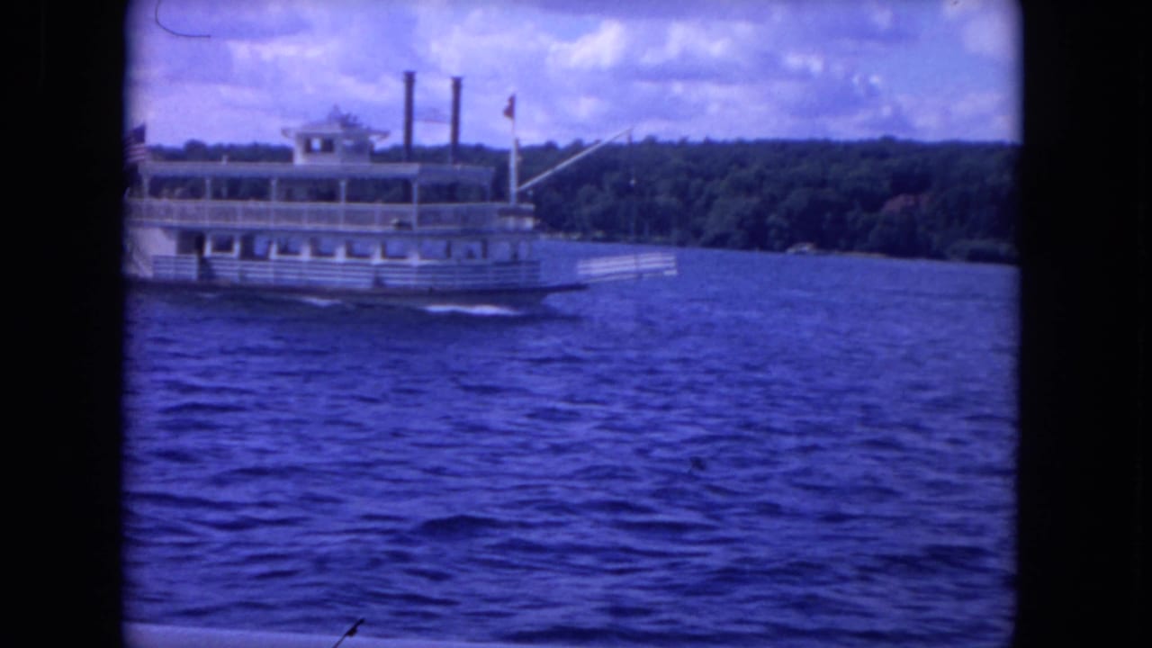 White riverboat on blue water with wooded shore and cloudy sky behind it.