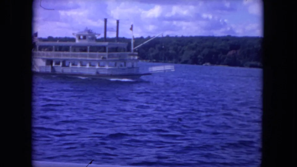 1972 8mm Mississippi River view of a white paddlewheel-style riverboat moving across blue water near a wooded shore.
