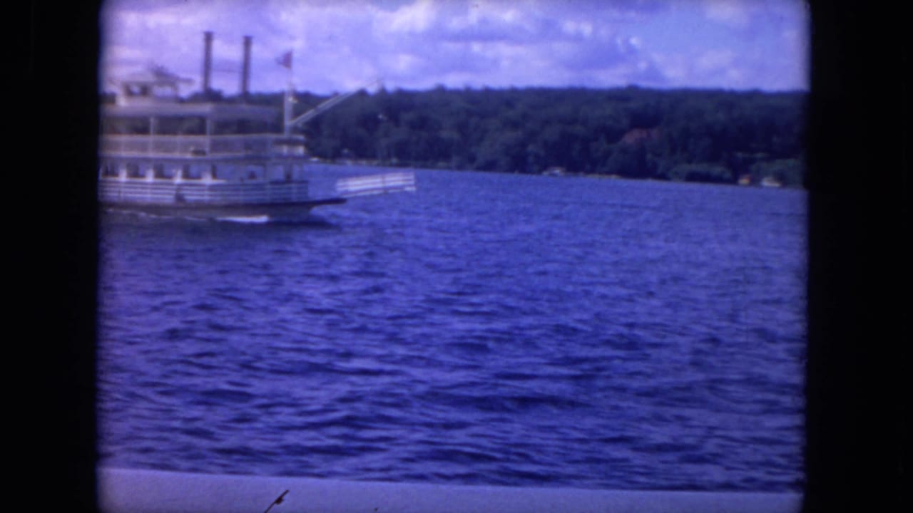 Riverboat partly at left on blue water with tree-lined shore beyond.
