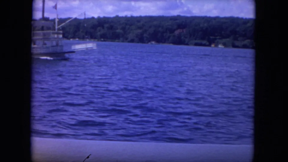 1972 8mm Mississippi River view with the riverboat nearly exiting frame, leaving rippled blue water and a green far bank.