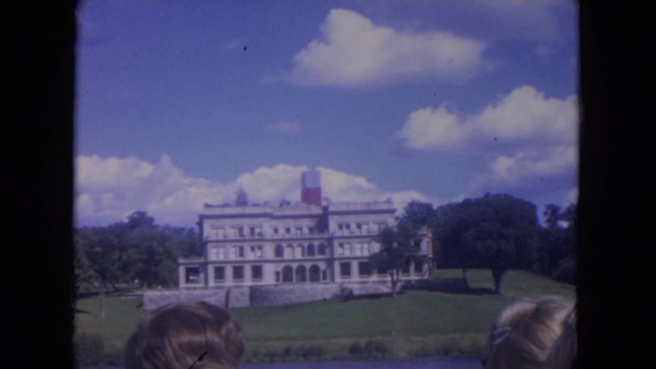 White mansion on a grassy hill seen from a riverboat with spectators at the bottom edge.