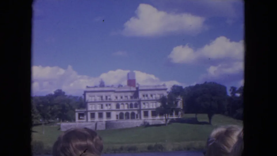1972 8mm Mississippi River view of a large white mansion on a green bank, framed from a moving boat with partial heads in the foreground.