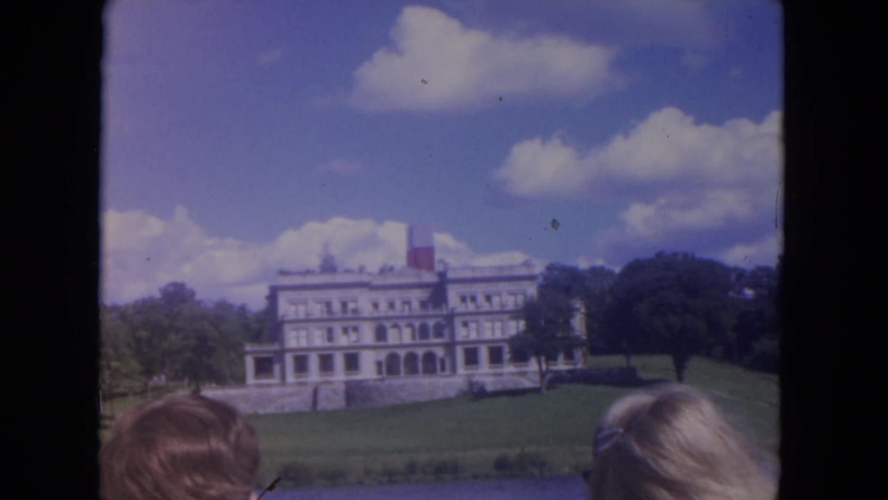 The riverboat passes a white mansion on a landscaped riverbank, with trees, clouds, and partial passengers visible at the frame edge.