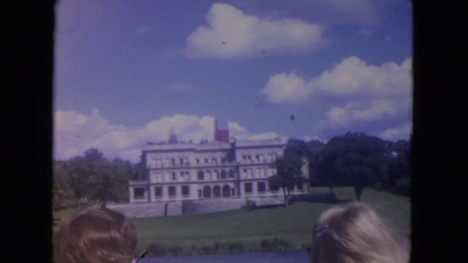 The riverboat passes a white mansion on a landscaped riverbank, with trees, clouds, and partial passengers visible at the frame edge.