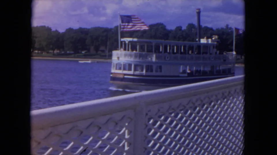 A white passenger riverboat with an American flag moves along the Mississippi River behind a white lattice railing in the foreground.
