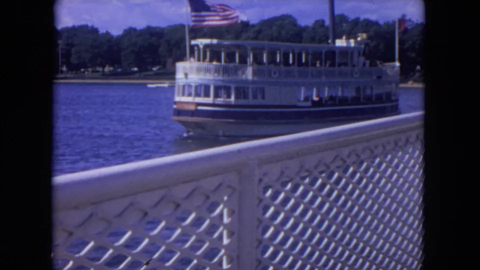 A vintage-looking white riverboat cruises past on the river, seen over a decorative lattice railing with trees lining the far bank.
