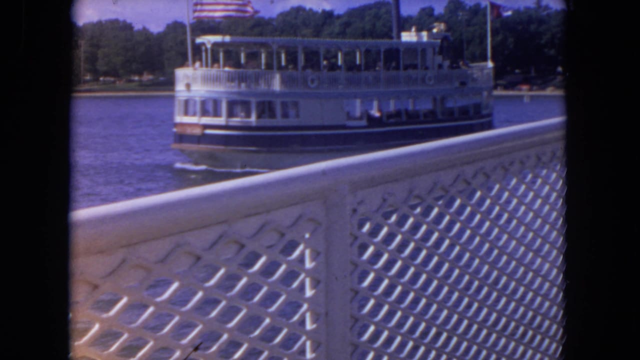 White riverboat continuing past a foreground lattice railing with shoreline trees behind it.