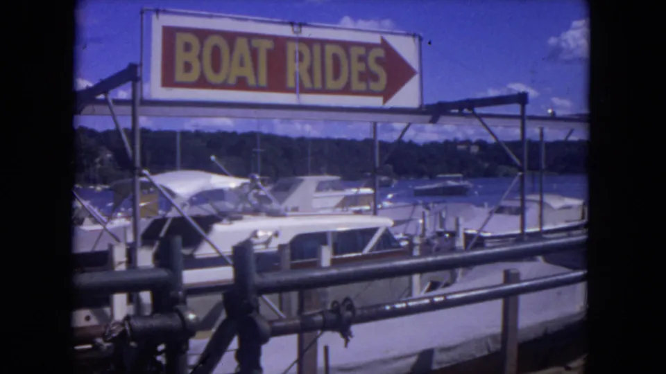 A red and yellow BOAT RIDES sign points across a marina area with boats, dock rails and river water below.