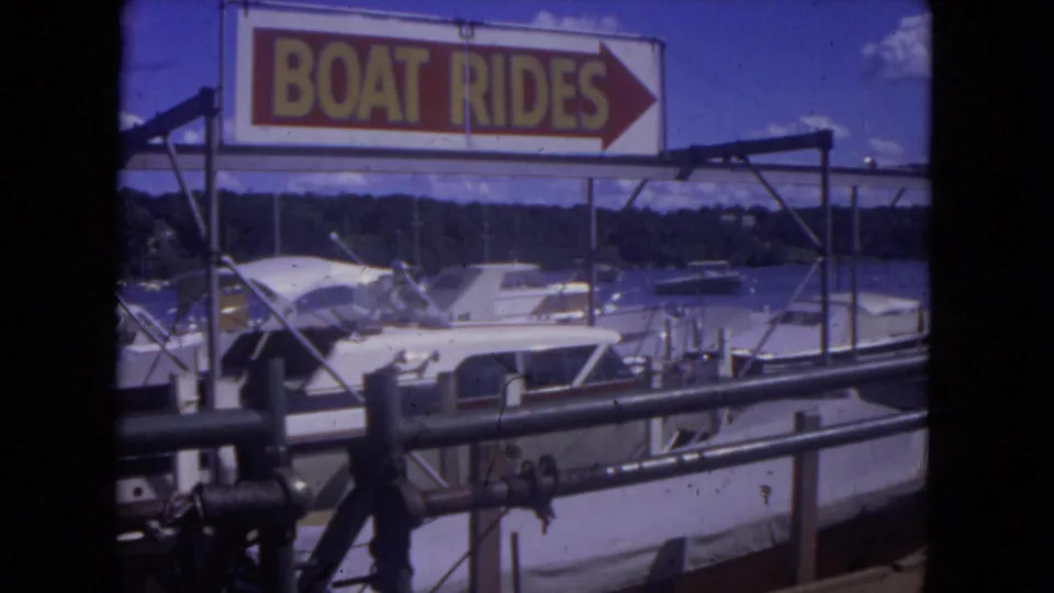 1972 Mississippi River boat dock with a bold BOAT RIDES sign, white cabin boats, foreground railings, and distant wooded shore.