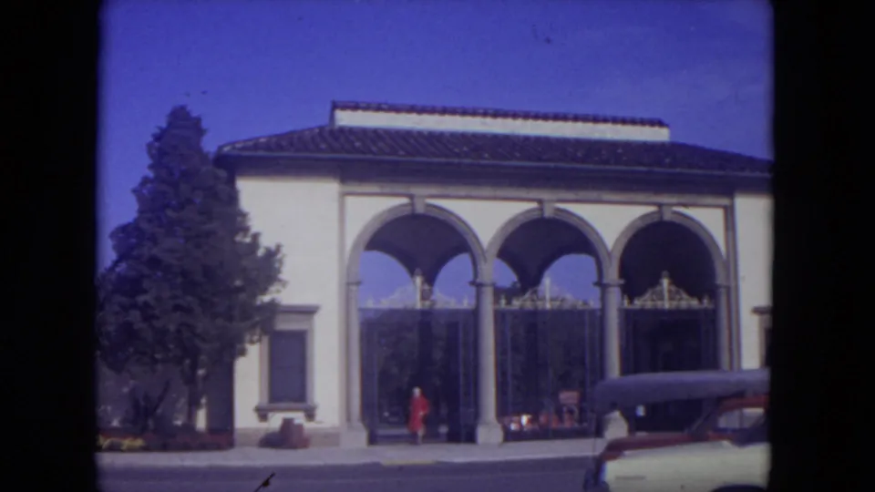 1972 travel scene near the Mississippi River area showing an arched building, tiled roof, a person in red, and a classic car at the curb.
