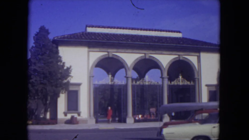 1972 Lincoln Park street view with a woman in red near a cream arched facade, iron gates, a tree, and a car carrying a roof load.